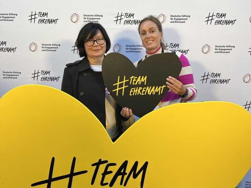 Zwei Personen halten ein großes gelbes Herz mit der Aufschrift „#Team“